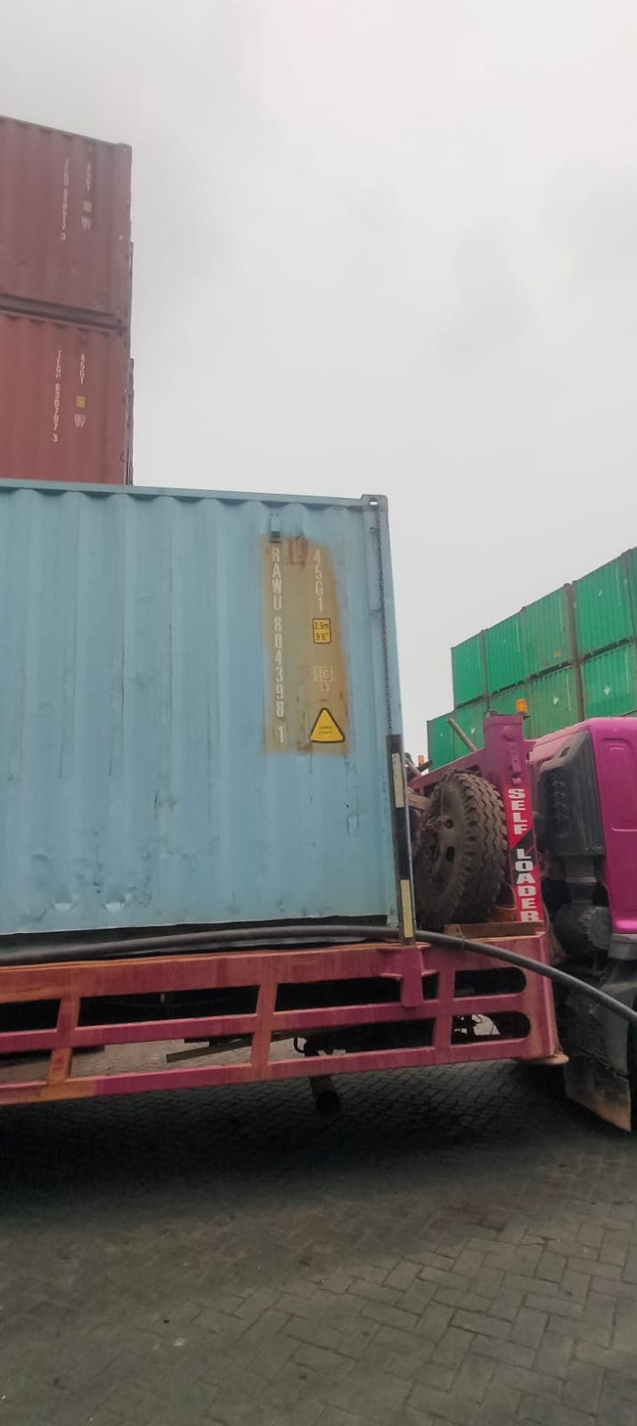 Kontainer biru muda berada di atas truk self loader di antara susunan kontainer